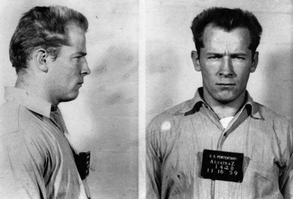 Black and white mugshot of Whitey Bulger. The left image shows a side profile, and the right image shows a front view. He is wearing a collared shirt and holding a board with "Alcatraz" and numbers readable. The expression is serious.