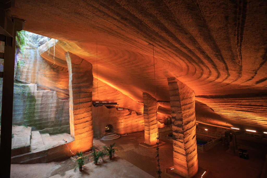 The image shows an underground cave with smooth, carved stone walls and columns. Warm lighting highlights the textured ceiling and walls. Some green plants are visible, and there are a few small seating areas.