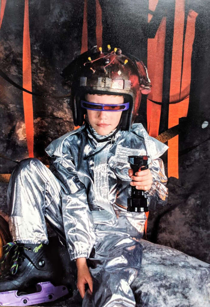 A child wearing a silver spacesuit costume and futuristic helmet sits on a rock. They have futuristic goggles and hold a toy device, resembling a sci-fi character. The background has dark tones with orange accents.