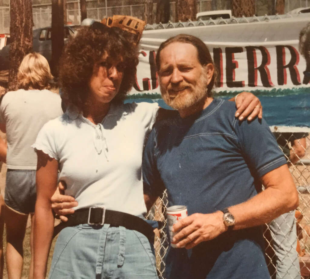 A woman with curly hair playfully sticks out her tongue while posing with a bearded man holding a can of beer. They stand outdoors in front of a chain-link fence with people and a partially visible banner in the background.