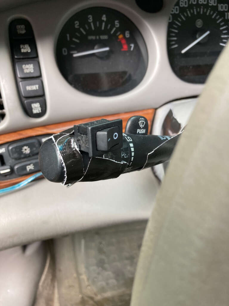 A car's interior with a close-up of the steering column. There is a piece of black tape wrapped around the turn signal lever, holding a small switch in place. The dashboard gauges are partially visible in the background.