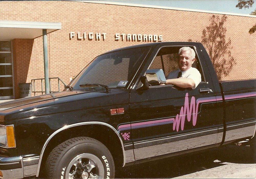 Man sitting in a black pickup truck with purple graphics, parked in front of a brick building labeled "Flight Standards.