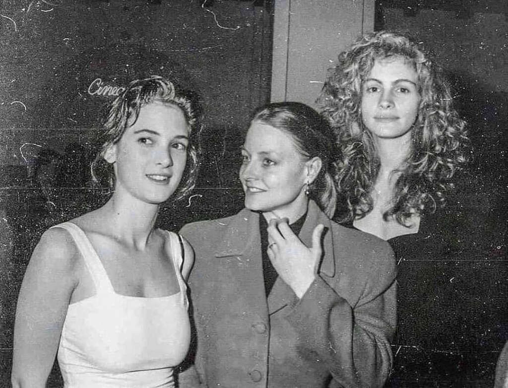 Three women pose together at an event. The one on the left wears a white dress, the middle woman wears a blazer, and the woman on the right has curly hair and wears a dark top. The background is dimly lit. The image is in black and white.