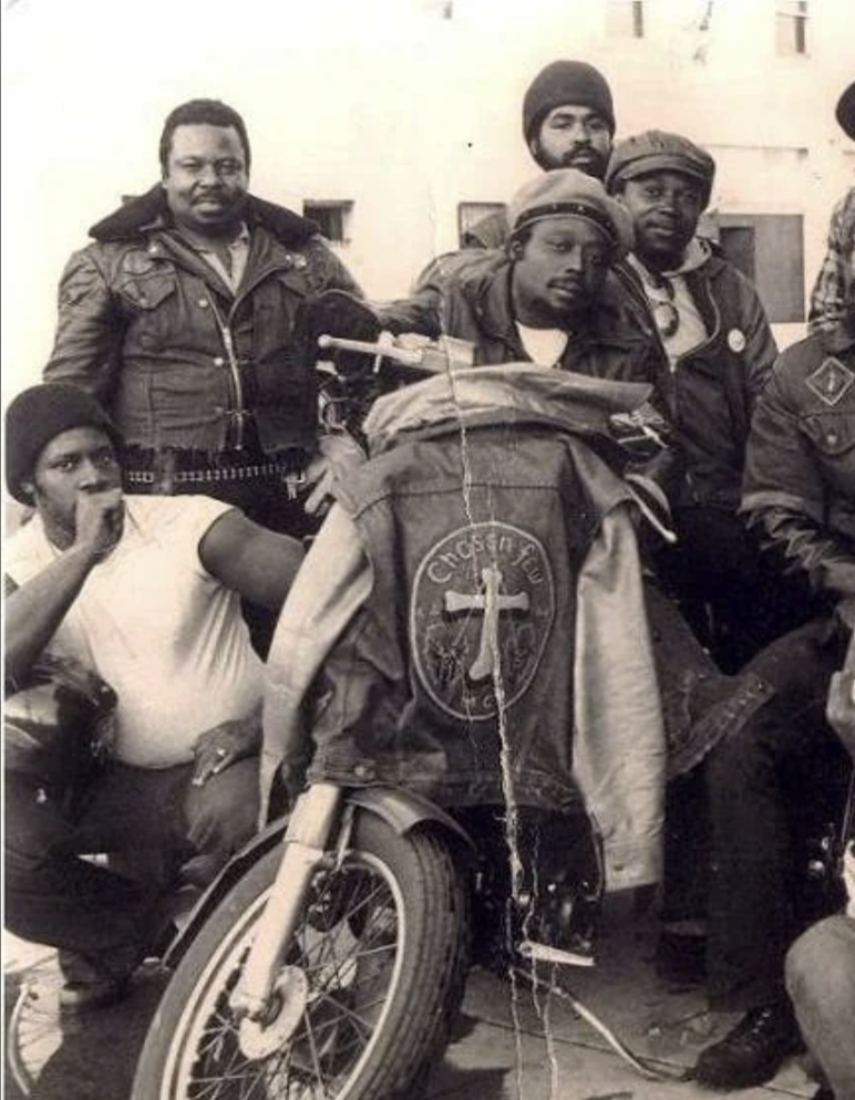 A vintage black-and-white photo shows a group of men posing with a motorcycle. They are wearing jackets, one featuring the patch "Chosen Few MC." The scene suggests camaraderie among motorcycle club members.