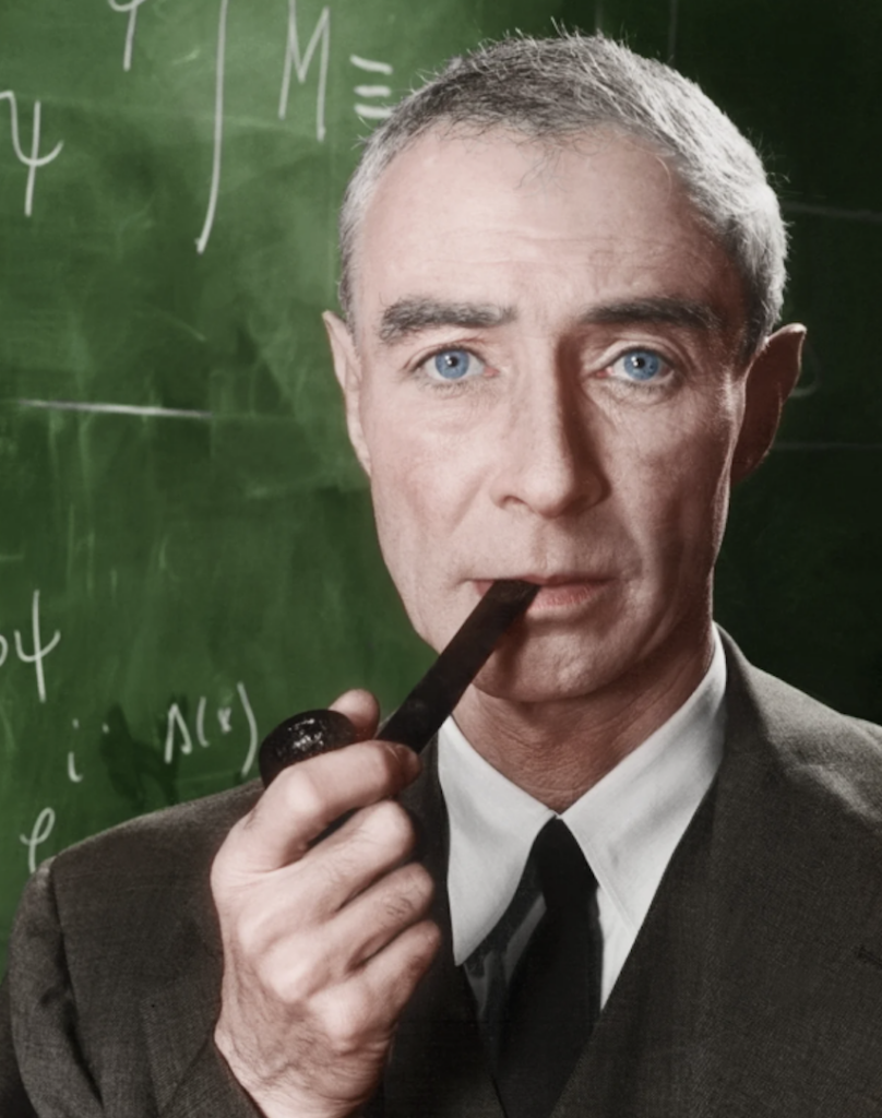 A person with short gray hair and blue eyes is holding a pipe in front of a chalkboard with mathematical equations. They are wearing a suit and tie.