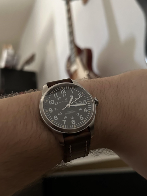 A wristwatch with a brown leather strap and a dark round face displaying both 12-hour and 24-hour times is shown on a person's wrist. In the blurred background, a guitar is visible against a plain wall.