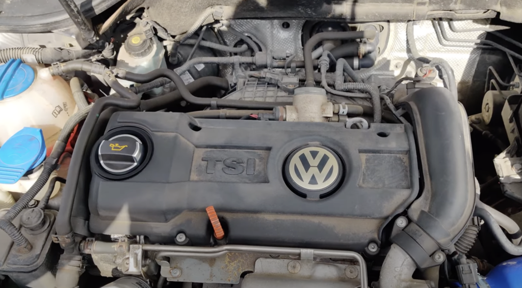 Engine compartment of a vehicle featuring a Volkswagen emblem and a TSI label. The engine cover has a visible oil cap with the oil can symbol. There's a partially visible coolant reservoir on the left with blue caps.