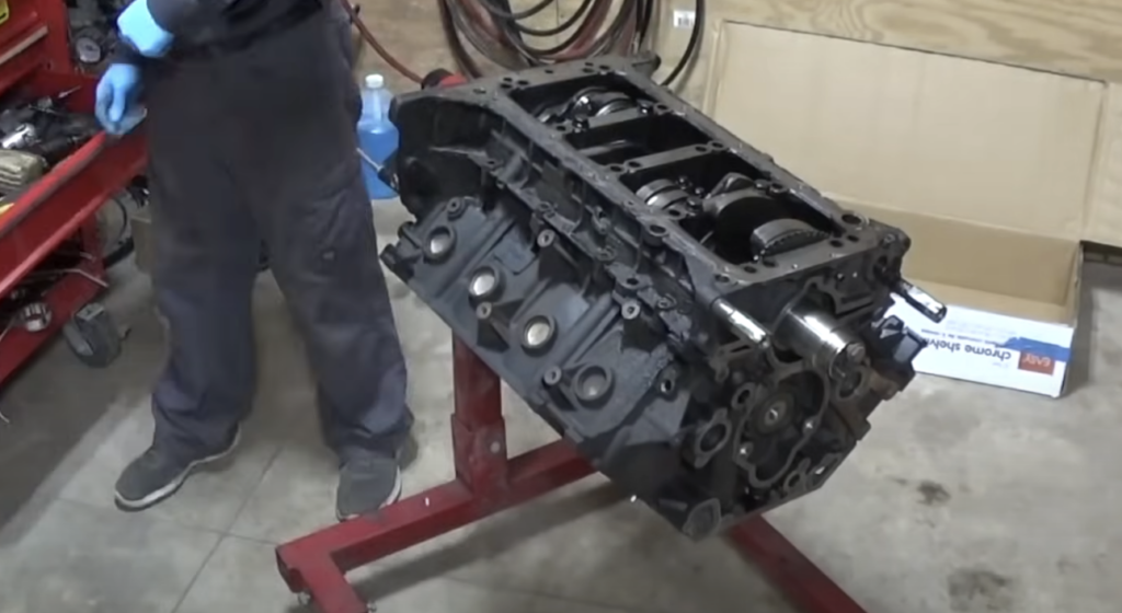 A person stands next to a disassembled engine block mounted on a red engine stand in a garage. Tools and a red toolbox are visible on the left. A cardboard box and various automotive parts are in the background.