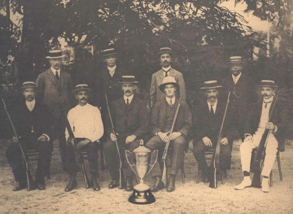 A vintage black and white photo of ten men posing outdoors with a large trophy. They are wearing suits, hats, and holding rifles. Some are seated while others stand behind them, with trees in the background.