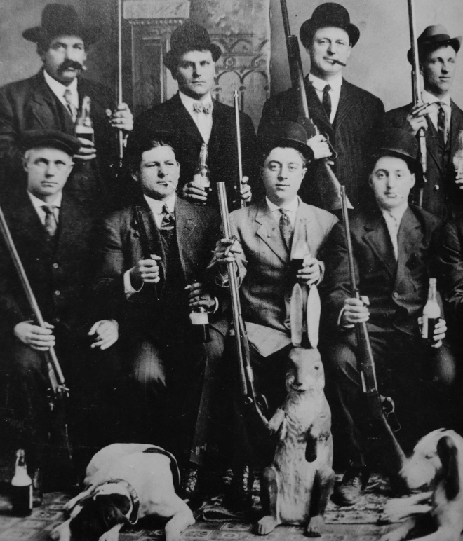 A black and white vintage photo of eight men posing with rifles and beverages. They are wearing suits and hats. In front, there is a large jackrabbit and two hunting dogs lying down. The backdrop is an ornate wall.