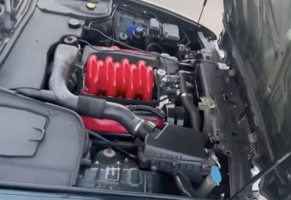 Car engine with a distinctive red intake manifold. The hood is open, revealing the various components like air filters, hoses, and fluid reservoirs. The engine bay is clean, and the red intake adds a pop of color.