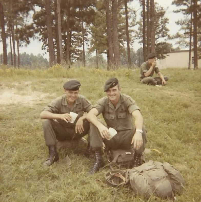 Two uniformed soldiers sit on grass, smiling and holding cups, with gear and a backpack nearby. Another soldier is in the background sitting and drinking. Tall pine trees and a building are visible under a cloudy sky.