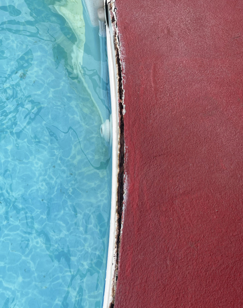 Close-up of a pool edge showing a worn and damaged red surface next to clear blue water. The edge is visibly cracked and peeling, highlighting a stark contrast between the vibrant blue and the aged red.