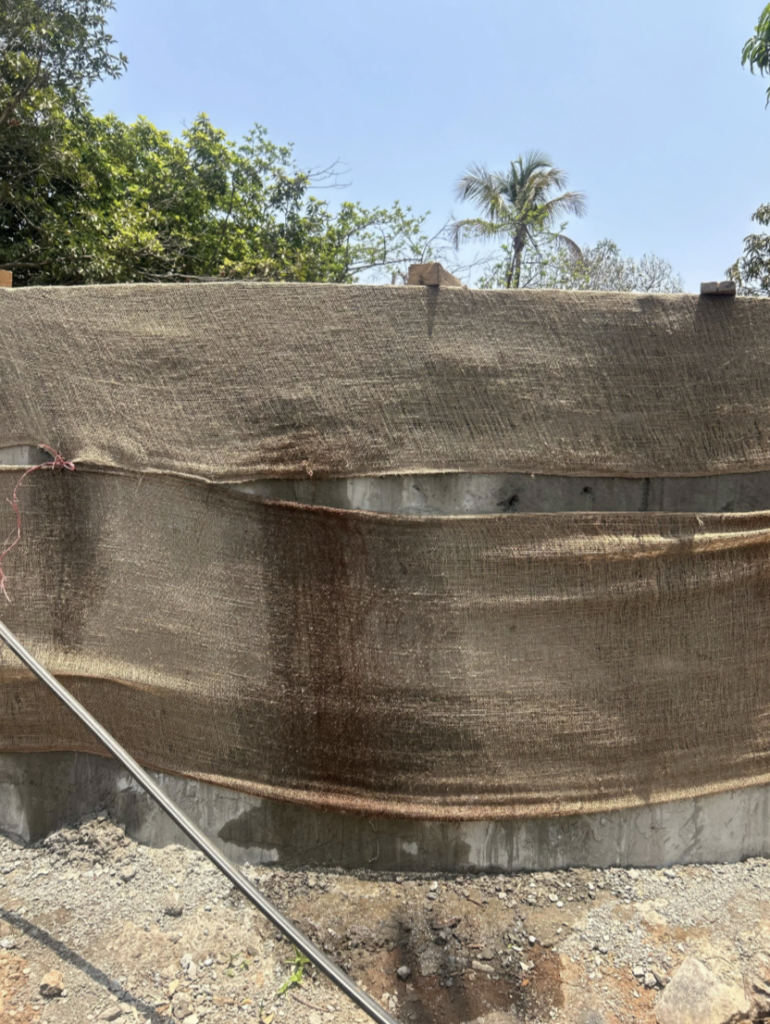 A construction site with a concrete wall partially covered by a brown tarp. There are metal rods protruding from the ground and a clear blue sky above. Trees and a palm tree are visible in the background.