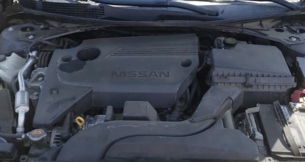 Close-up view of a Nissan car engine, showing various components including the engine cover with the Nissan logo, air filter housing, battery, and connecting wires. Some parts have light dust or wear.