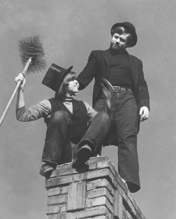 Two men in vintage attire are on a brick chimney. One is crouching, holding a chimney sweep brush and wearing a top hat. The other stands, wearing a bowler hat and holding the crouching man's hand. The sky is clear in the background.