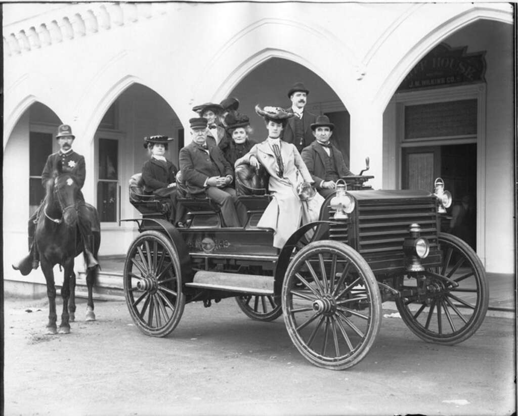 A vintage black and white photo of seven people in an early automobile with large wheels, parked on a street. A man on horseback stands nearby. The passengers and driver wear formal clothing, and an arched building facade is visible in the background.