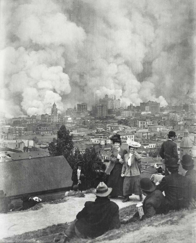 A historical black and white photo shows people sitting on a hill, observing a city engulfed in smoke and flames. The skyline is dominated by large plumes of smoke. The clothing suggests an early 20th-century setting.