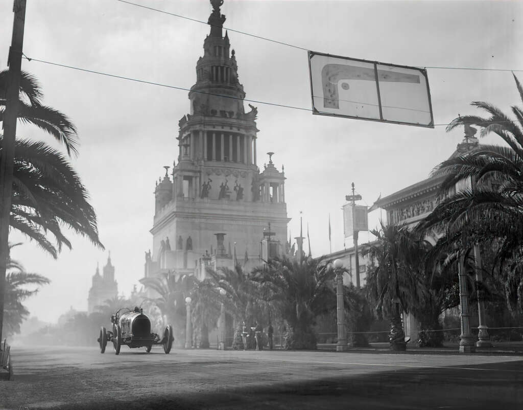 A vintage car races on a broad street lined with palm trees, passing a grand, ornate building with statues and a tower. The scene is misty, with another majestic structure visible in the background, under a cloudy sky.