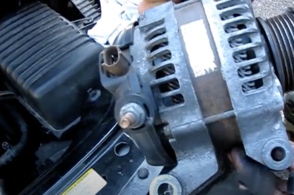 A person is holding an alternator part next to a car engine. The alternator has a silver, metallic exterior with vented sides and visible connection points. Nearby, the car's engine components and a black cover can be seen.