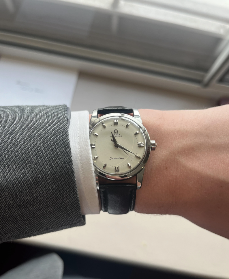 A person wearing a watch with a silver case, white dial, and black leather strap. The watch has a brand logo and "Seamaster" text on the face. The person is wearing a grey suit jacket and white shirt. A window and papers are blurred in the background.