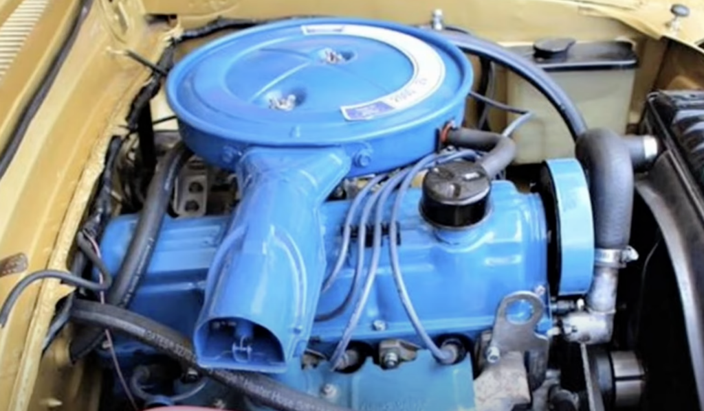 Close-up of a car engine in an open hood. The engine is painted bright blue with a circular air filter cover on top. Surrounding parts are black and metallic, with various tubes and wires extending around. The car's exterior is painted beige.