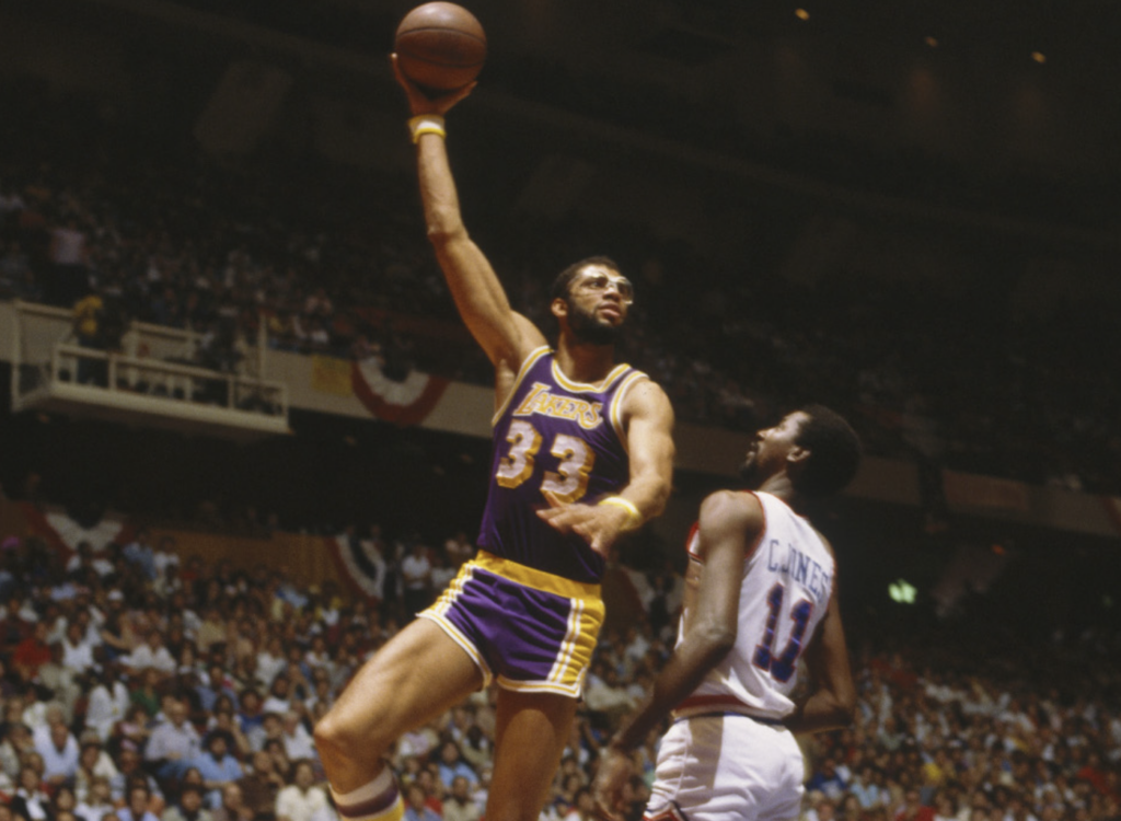 Basketball player in a purple and yellow Lakers uniform, number 33, jumps with his arm extended holding a basketball. An opposing player, number 13, in a white uniform watches. The arena is filled with spectators.