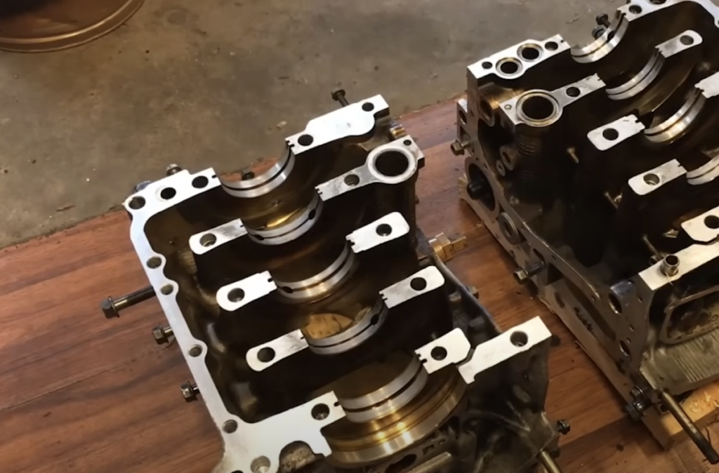 Two engine blocks are placed on a wooden workbench, showcasing their intricate internal components. The metal surfaces are polished, revealing the cylindrical spaces and bolt holes, indicative of mechanical work or repair.