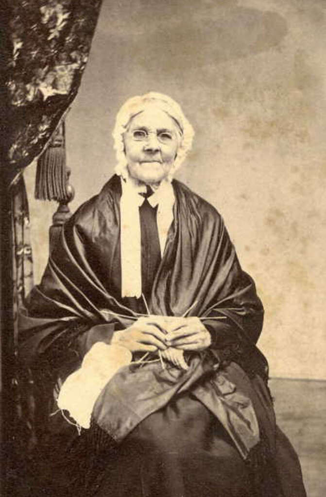 An elderly woman wearing glasses and a bonnet sits in a chair. She is draped in a dark shawl and holds needlework in her hands. A partially visible curtain is behind her. The photo is sepia-toned.