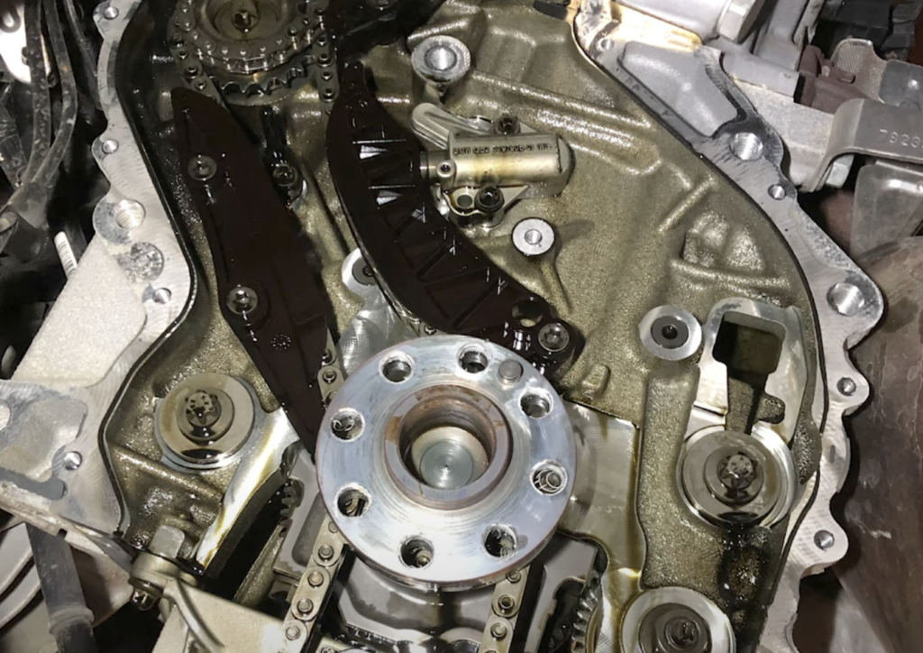 Close-up of an engine part with visible gears, chains, and metal components. The area appears oily, and bolts are present, indicating mechanical complexity. The image reveals intricate details of the internal mechanics.