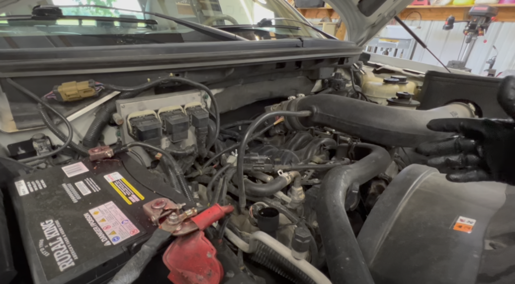 The image shows a close-up view of a vehicle's engine compartment. Various components, wiring, and hoses are visible. A gloved hand is gesturing towards the engine. A car battery with labels is on the left side.