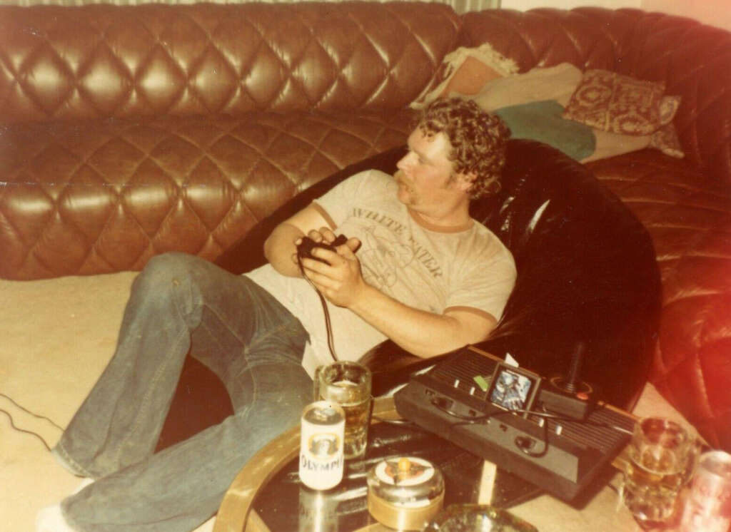 A man with curly hair and a mustache is sitting in a black bean bag chair, playing a video game with a joystick. He's in a room with a brown quilted sofa. Cans and glasses are on a small table in front of him.