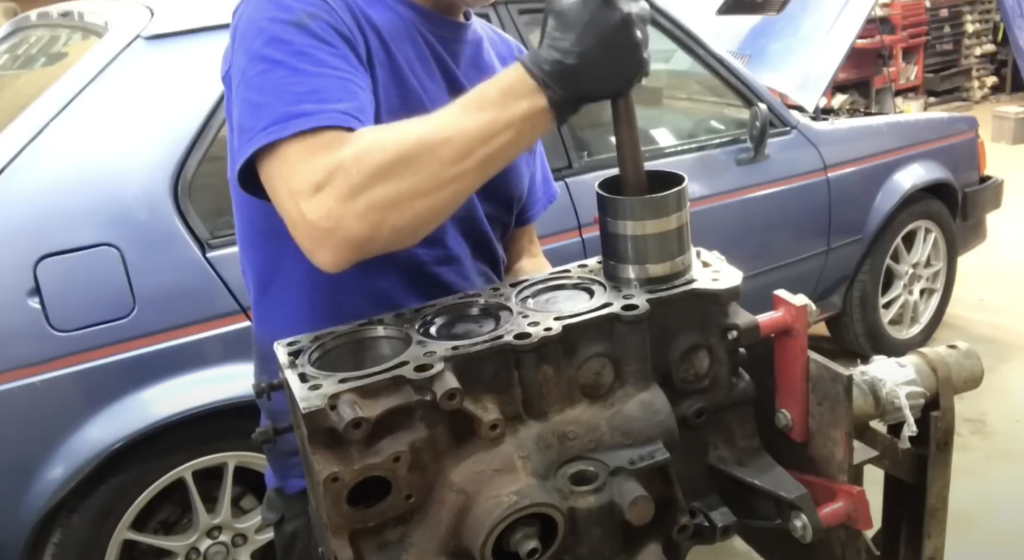 Person in a blue shirt works on a car engine block in a garage. They are using a tool on the engine while standing next to a blue car with silver rims. The background features shelves and automotive supplies.