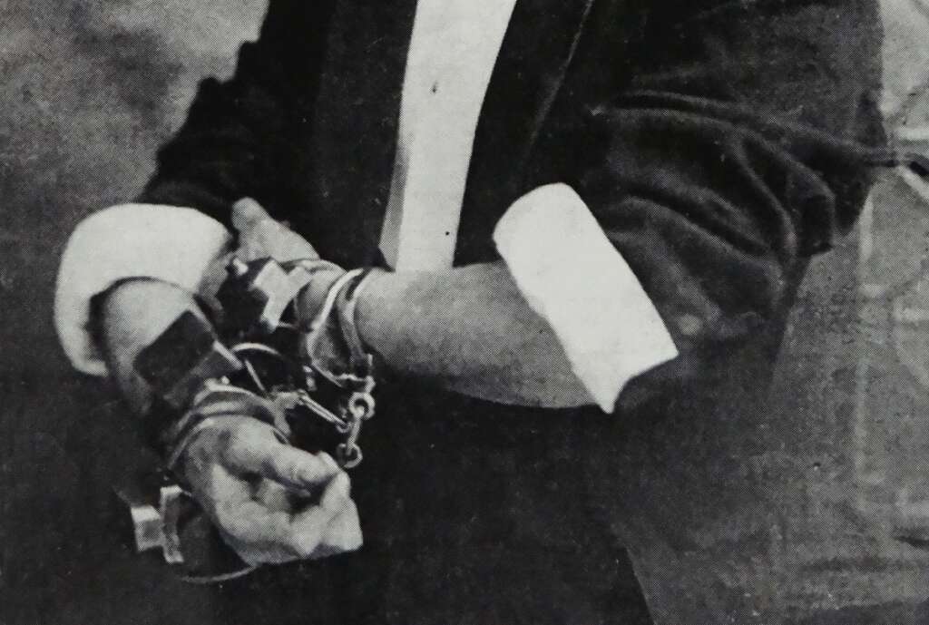 A person in a suit is shown with their arms bound by multiple handcuffs. The sleeves of the suit are rolled up slightly, revealing the cuffs tightly wrapped around the wrists against a blurred background.