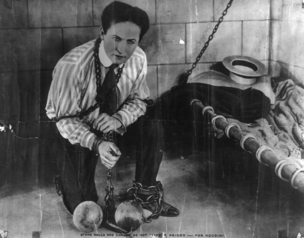 A person in a striped shirt and tie is kneeling, wrapped in chains and shackles, next to a bed with a large metal ring on the floor. The setting appears to be a cell. Text below reads, "Stone walls and chains do not make a prison for Houdini.