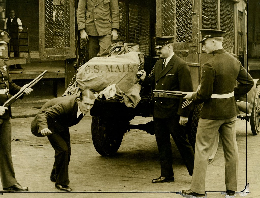 Harry Houdini Escapes - Harry Houdini in a suit is bending down in front of an armed inspector as a mail truck is being unloaded. Two postal workers stand nearby with another armed inspector. A large U.S. Mail sack is being handled near the truck.