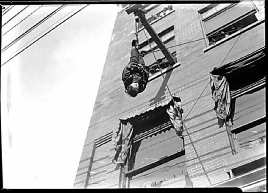 A person is hanging upside down from a pole attached to a multi-story brick building. The building has several windows, some with awnings. The scene is black and white, conveying a vintage or historical atmosphere.