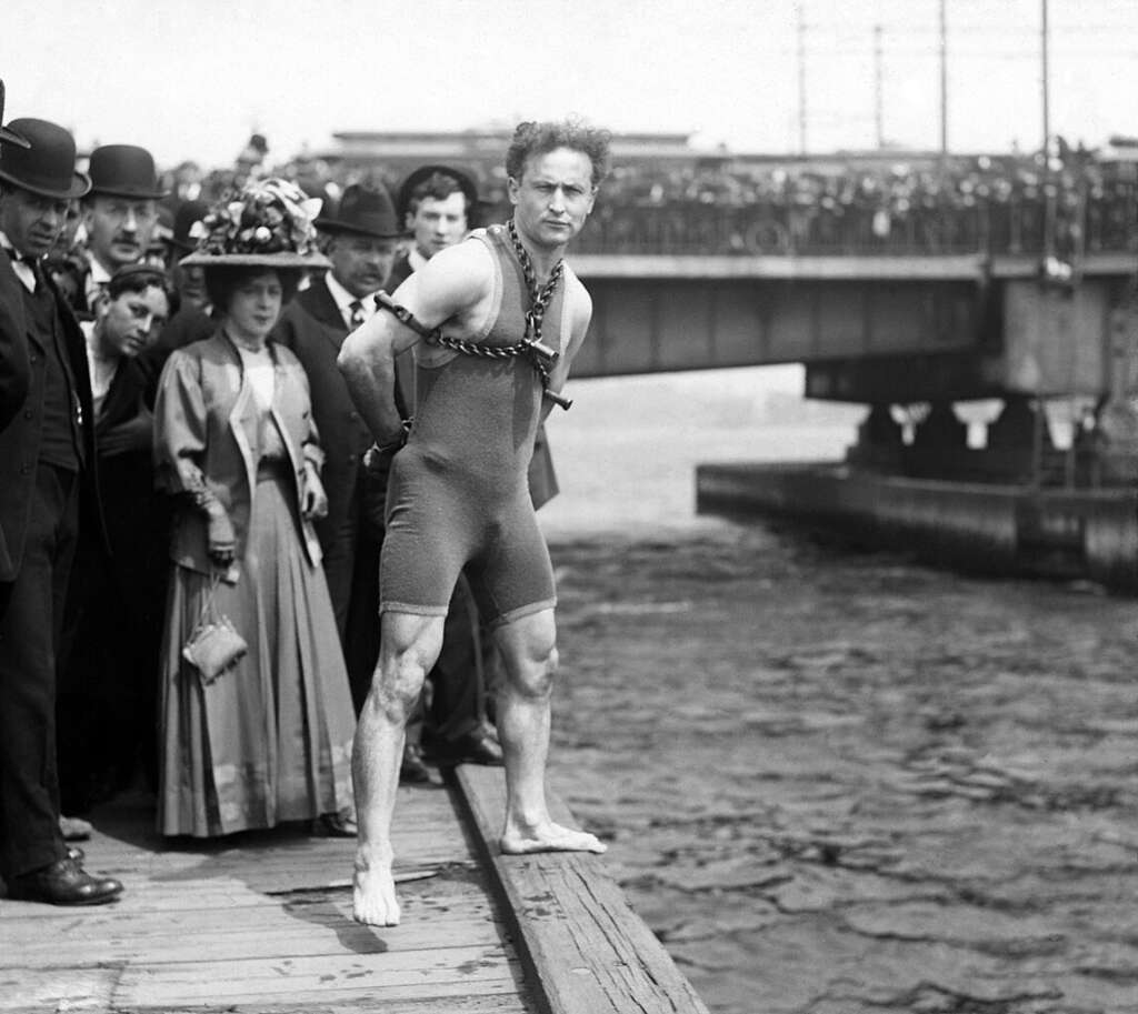 A man in a sleeveless suit stands barefoot on a dock, bound with chains and locks, preparing for a stunt jump into the water. Onlookers in early 20th-century attire watch from the dock and a bridge in the background.