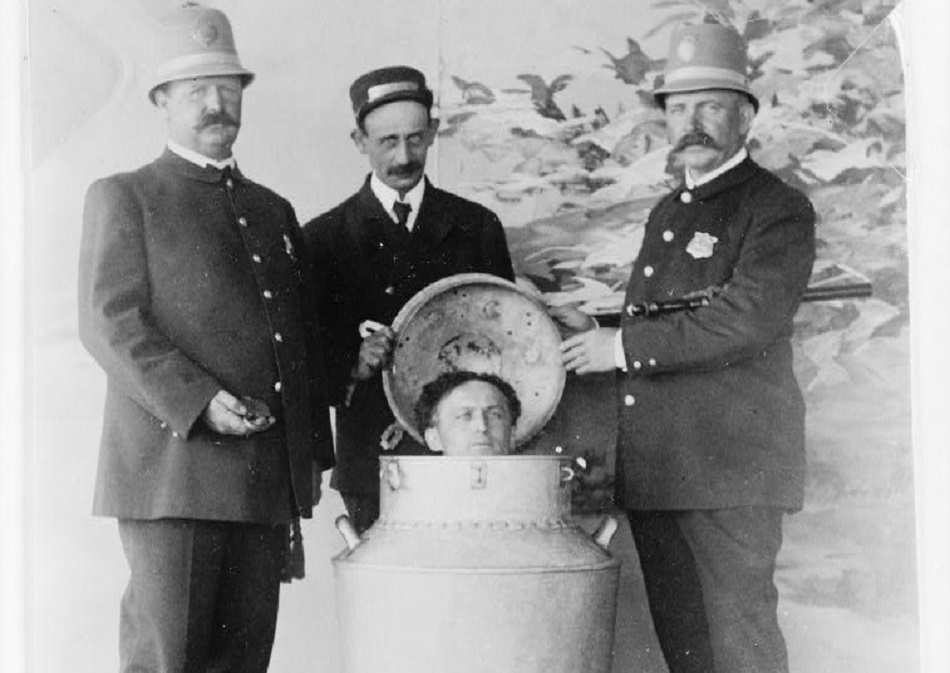 Three men in uniforms stand around a large metal canister. One lid is open, revealing a fourth man's head inside the canister. The setting appears to be an old, possibly staged, photograph with a painted backdrop.