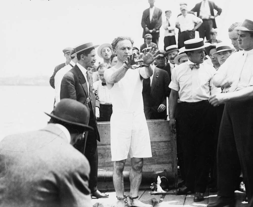 A man in a white outfit with his hands shackled stands on a dock, surrounded by a crowd of onlookers in early 20th-century attire, including straw hats and suits. The scene appears to be a public spectacle or performance.