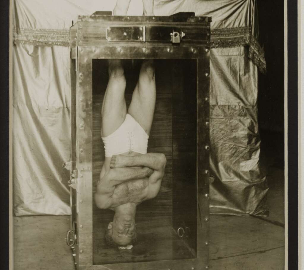 A man in white shorts is submerged upside down in a transparent, water-filled tank, secured with locks. A draped background surrounds the tank, highlighting the dramatic nature of the situation.