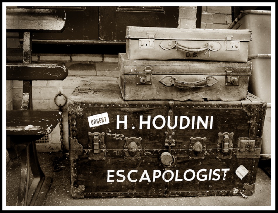 A sepia-toned image of two vintage suitcases stacked on an old trunk. The trunk has labels, including "URGENT" and "H. HOUDINI ESCAPOLOGIST." The backdrop shows a bench and a brick wall.