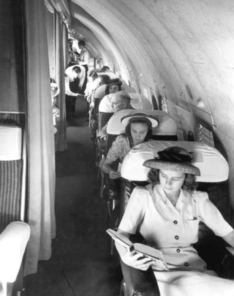 Black and white photo of passengers seated in a narrow airplane cabin from the past. Women in front are wearing dresses and hats, one reading a book. Flight attendant seen in the aisle mid-cabin.