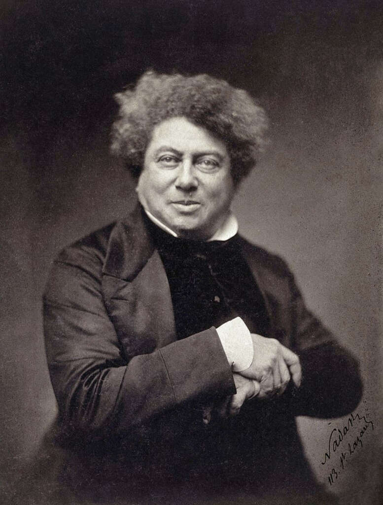 A sepia-toned portrait of a man with curly hair, dressed in a dark suit with a high-collared shirt. He is seated, hands resting on a cane. His expression is neutral, and the background is a plain, blurred shade. A signature is visible in the lower right corner.