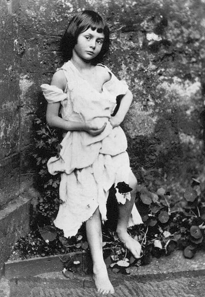 Black and white photo of a young girl standing barefoot against a rough stone wall. She is wearing a tattered, loose-fitting dress and has a thoughtful expression, with one foot crossed over the other amidst scattered foliage.