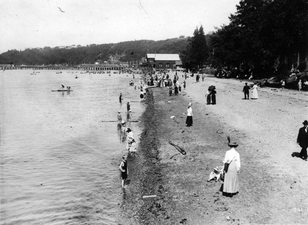 Black and white photo of a historical beach scene with people in early 20th-century clothing. Men, women, and children are near the shoreline, some playing in the water. Trees and a building are in the background.