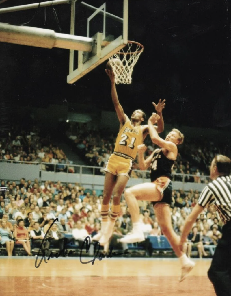 A basketball player in a yellow uniform jumps to make a layup while being closely guarded by a player in a black uniform. The game is in an indoor arena with spectators watching intently. An official in a striped shirt observes nearby.