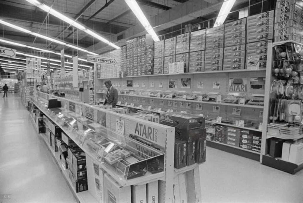 Black and white photo of an electronics store aisle from the 1980s. Shelves are stocked with Atari products, including game consoles and software. A person is browsing items alone in the spacious, brightly lit store.