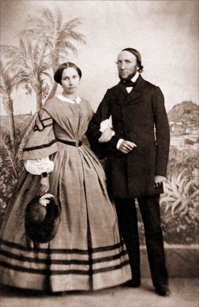 A vintage photograph of a man and woman in formal 19th-century attire. The woman wears a long dress and holds a small hat. The man is dressed in a dark suit. They stand against a painted backdrop depicting a landscape with palm trees.