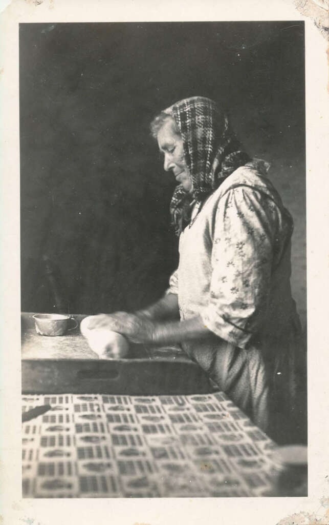 An elderly woman wearing a headscarf and a patterned blouse kneads dough on a table. A small bowl is on the table beside her. The scene is in black and white, with a patterned cloth in the foreground.